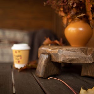 Puede incluir: Una mesa de pedestal de madera r&uacute;stica con una taza de caf&eacute; y un jarr&oacute;n con flores secas. El pedestal es de madera marr&oacute;n oscuro y tiene una superficie rugosa y texturizada. La taza de caf&eacute; es amarilla con tapa blanca.