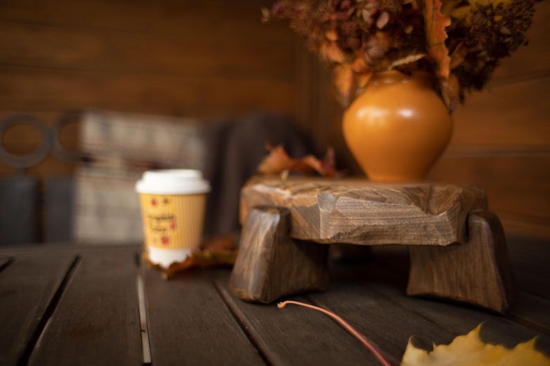 Puede incluir: Una mesa de pedestal de madera r&uacute;stica con una taza de caf&eacute; y un jarr&oacute;n con flores secas. El pedestal es de madera marr&oacute;n oscuro y tiene una superficie rugosa y texturizada. La taza de caf&eacute; es amarilla con tapa blanca.
