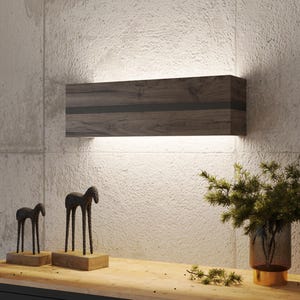 Op de afbeelding: Een moderne wandlamp met een donkere houten afwerking en een zwarte accentstreep. Het licht is gemonteerd op een betonnen muur met textuur. Twee bronzen paardensculpturen staan op een houten oppervlak.