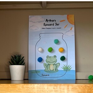 Op de afbeelding: Een beloningskaart met een kikkerillustratie in een pot, met de tekst "Arthurs Reward Jar". De kaart heeft kleurrijke pompons en is ontworpen om punten te verzamelen voor beloningen. Links staat een kleine pot met een vetplant.