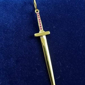 Op de afbeelding: Een goudkleurige hanger in de vorm van een zwaard. Het handvat is versierd met kleine, roze edelstenen. De hanger heeft een lus aan de bovenkant voor een ketting. Het is geplaatst tegen een donkerblauwe achtergrond.