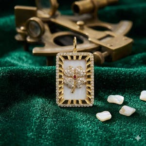 May include: Gold pendant with a rectangular frame encasing a white background and a four-leaf clover design. The clover is adorned with small diamonds and a central red gemstone. The pendant hangs from a gold loop.