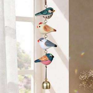 Puede incluir: Móvil decorativo colgante con cuatro figuras de pájaros pintados en azul, blanco, naranja y rosa. Los pájaros están conectados por anillos metálicos y suspendidos de un cordón marrón. Una pequeña campana dorada cuelga en la parte inferior.