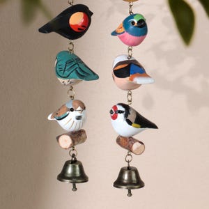 May include: Hand-painted wooden bird wind chimes. Each chime features several colorful birds hanging from a chain, with a small bell at the bottom. The birds are perched on small wooden branches. The birds are painted in various colors, including black, blue, and white.