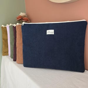 Peut inclure: Plusieurs pochettes à fermeture éclair en velours côtelé de différentes couleurs, dont bleu marine, marron et violet. La pochette bleu marine porte une étiquette blanche avec le mot "Maarja". Les pochettes sont présentées sur une surface blanche.