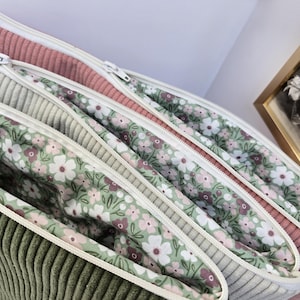 Peut inclure: Gros plan sur trois pochettes à fermeture éclair en velours côtelé vert, rose et crème. Les pochettes ont des fermetures éclair blanches et des doublures à motifs floraux avec des fleurs roses et blanches sur fond vert. Le velours côtelé a une texture côtelée verticale.