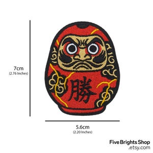 K&ouml;nnte beinhalten: Ein roter Daruma-Puppen-Aufn&auml;her, 7 cm hoch und 5,6 cm breit, mit einem schwarzen und goldenen Design. Der Aufn&auml;her zeigt japanische Schriftzeichen und stammt von Five Brights Shop.