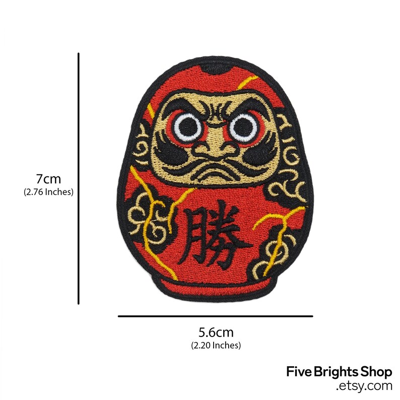 K&ouml;nnte beinhalten: Ein roter Daruma-Puppen-Aufn&auml;her, 7 cm hoch und 5,6 cm breit, mit einem schwarzen und goldenen Design. Der Aufn&auml;her zeigt japanische Schriftzeichen und stammt von Five Brights Shop.