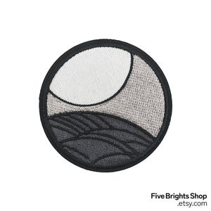 May include: Embroidered patch featuring a stylized landscape within a circle. The design includes a white crescent shape, gray textured area, and dark gray wavy lines, all outlined in black. The patch is likely for clothing or accessories.