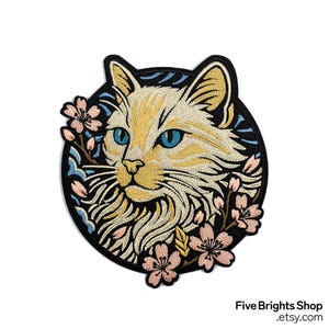 May include: Embroidered patch featuring a cat's face with blue eyes, surrounded by pink cherry blossoms and blue accents, set against a black background. The cat has yellow and cream-colored fur. The patch is circular in shape.