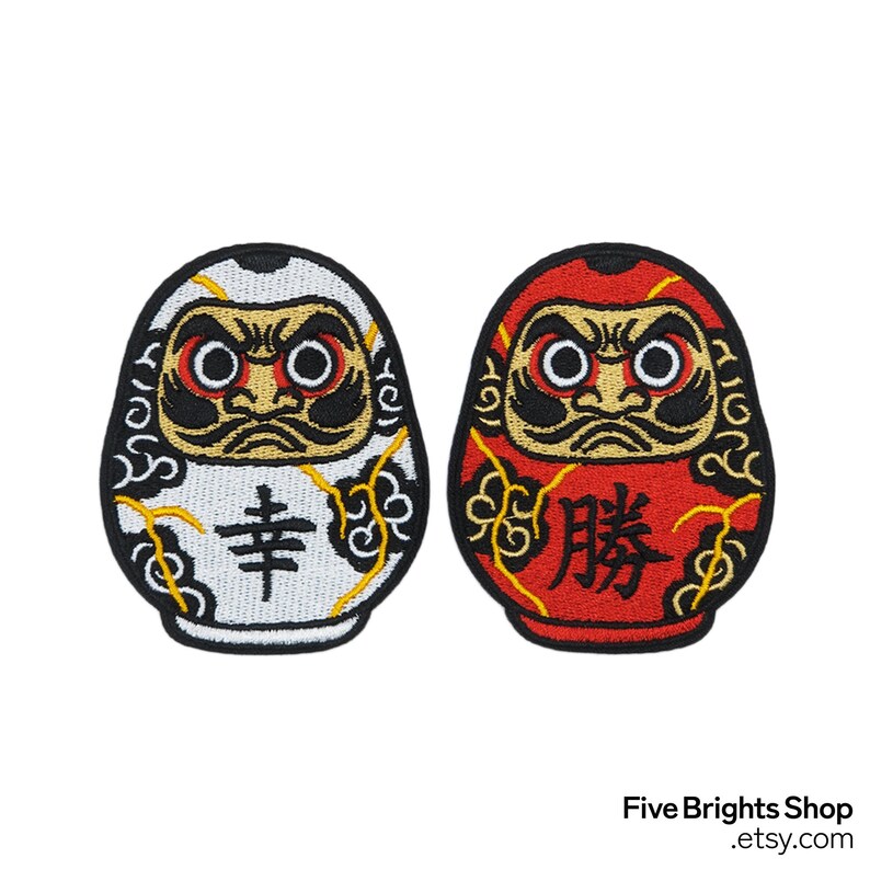 K&ouml;nnte beinhalten: Zwei Daruma-Puppen-Patches, einer wei&szlig; und einer rot. Die wei&szlig;e Puppe hat das japanische Zeichen f&uuml;r "Gl&uuml;ck", und die rote Puppe hat das Zeichen f&uuml;r "Sieg". Beide haben schwarze Details und goldene Akzente.