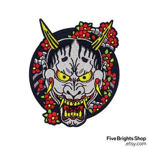 Può includere: Toppa ricamata raffigurante una maschera Oni grigia con occhi e corna gialle, bocca rossa e zanne gialle. La maschera è circondata da fiori rossi e foglie verdi su uno sfondo circolare nero. Il testo "Five Brights Shop .etsy.com" è in basso.