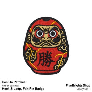 Può includere: Toppa termoadesiva a forma di bambola Daruma rossa con dettagli neri e dorati. La toppa presenta un design dettagliato della tradizionale bambola giapponese, con il carattere giapponese per "vittoria" ricamato in rosso.