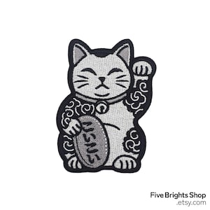Puede incluir: Parche bordado con un Maneki-neko, o gato de la suerte japonés, en tonos grises y negros. El gato está representado con una pata levantada y sosteniendo una moneda. El parche tiene un borde negro y remolinos decorativos.