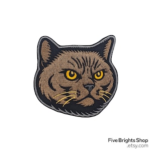 May include: Embroidered patch featuring a brown cat face with yellow eyes and whiskers. The cat has a black outline and a gray border. The patch is a detailed, close-up design of a cat's face.