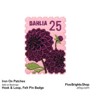 以下が含まれることがあります： 紫色のダリアの花のデザインが施されたピンク色のアイロン接着パッチ。パッチには切手のような縁取りがあり、「DAHLIA」の文字と数字の「25」が描かれています。緑の葉とつぼみも含まれています。