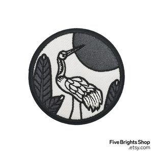 Könnte beinhalten: Runder, gestickter Aufnäher mit einem Kranich in minimalistischem Design. Der Kranich ist weiß mit schwarzen Umrissen, vor einem weißen Hintergrund mit einem dunkelgrauen Mond und schwarzem Laub. Der Aufnäher hat einen schwarzen Rand.