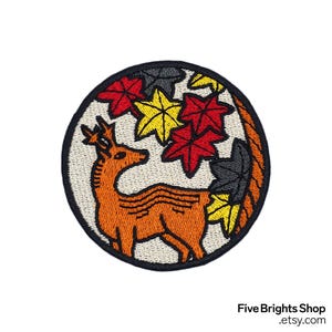 以下が含まれることがあります： オレンジ色の鹿と、赤、黄、グレーの葉に囲まれた、スタイリッシュな角を持つ刺繍パッチ。パッチは黒い縁取りとクリーム色の背景です。「Five Brights Shop .etsy.com」の文字が右下にあります。