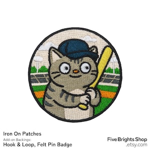 May include: Round iron-on patch featuring a cartoon cat in a baseball cap holding a yellow bat. The cat is gray with black stripes, set against a baseball field background. The patch has a stitched border.