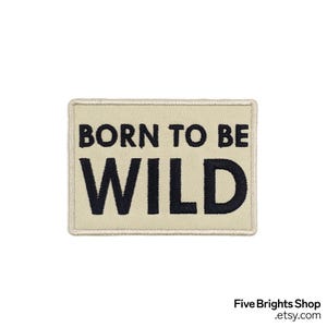 Può includere: Una toppa rettangolare con le parole "BORN TO BE WILD" in lettere nere su sfondo beige. La toppa ha un bordo cucito, adatta per abbigliamento o accessori.