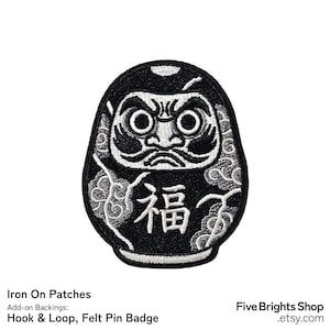 Peut inclure: Un écusson brodé noir et blanc représentant une poupée Daruma. L'écusson présente un visage stylisé avec de grands yeux et des sourcils épais au-dessus du caractère kanji japonais pour la bonne fortune, entouré de motifs de nuages tourbillonnants.