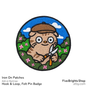 Könnte beinhalten: Runder Aufnäher zum Aufbügeln mit einer Cartoon-Katze, die einen braunen Hut trägt und eine Gartenschere hält. Die Katze ist von grünen Büschen, rosa Blumen und einem blauen Himmel mit Wolken umgeben. Der Aufnäher trägt den Text "Iron On Patches" und "Hook & Loop, Felt Pin Badge".