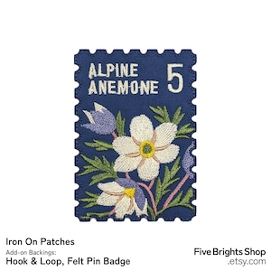 Könnte beinhalten: Marineblauer, gestickter Aufnäher in Form einer Briefmarke. Er zeigt die Worte "Alpine Anemone" und die Zahl "5" in Weiß. Der Aufnäher zeigt weiße und lila Blumen mit grünen Blättern.