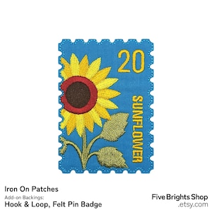 Op de afbeelding: Geborduurde patch in de vorm van een postzegel met een zonnebloem. De patch is blauw met een gele zonnebloem, groene bladeren en het nummer 20. Het woord "SUNFLOWER" staat verticaal gedrukt.