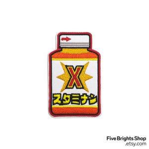 May include: Embroidered patch in the shape of a medicine bottle. The patch has a white background with a yellow and red border. The design includes a yellow 'X' and Japanese text.