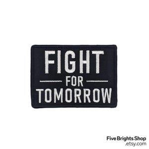 Könnte beinhalten: Ein rechteckiger schwarzer Aufnäher mit weiß gesticktem Text, der „FIGHT FOR TOMORROW“ lautet. Der Aufnäher hat einen weißen Rand und ist vor einem weißen Hintergrund platziert.