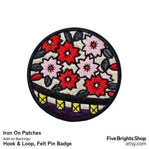 Op de afbeelding: Een ronde geborduurde patch met een tros rode en lichtroze kersenbloesems met donkergrijze bladeren op een crèmekleurige achtergrond. Het onderste gedeelte toont een donkerpaarse rand met een geel rasterpatroon en een zwarte rand.