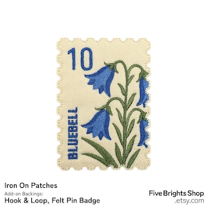 Pode incluir: Remendo bordado em forma de selo postal com flores de jacinto-azul. O remendo tem um fundo creme com uma borda ondulada. A palavra "BLUEBELL" e o número "10" são impressos no remendo.