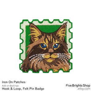 Puede incluir: Parche bordado con la cara de un gato con ojos azules, pelaje marrón y detalles amarillos, enmarcado en un diseño de sello postal verde. El parche está etiquetado como "Iron On Patches" e incluye opciones de respaldo de velcro y pin de fieltro.