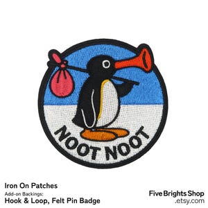 May include: Embroidered patch featuring a cartoon penguin with a red beak and a bag. The penguin is black and white, standing on a white base, with a blue and black circular border. The words "NOOT NOOT" are embroidered below the penguin.