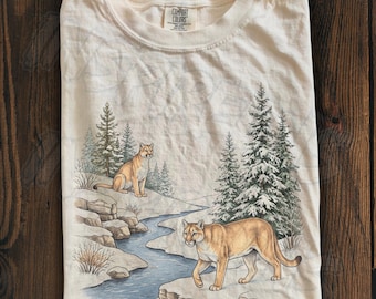 May include: A cream-colored t-shirt featuring a graphic print of two mountain lions in a snowy landscape with pine trees and a winding stream. The shirt has a crew neckline and a Comfort Colors brand label printed on the inside collar.