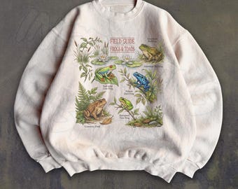 May include: A cream-colored crewneck sweatshirt featuring a graphic print titled Field Guide to Frogs & Toads. The design includes illustrations of various frogs, lily pads, and foliage with labels for American Bullfrog, Red-eyed Tree Frog, Poison Dart Frog, and European Tree Frog.