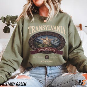 Puede incluir: Una sudadera de cuello redondo color verde oliva con un estampado gr&aacute;fico de un castillo en las monta&ntilde;as. El texto dice TRANSYLVANIA, Castle of Dracula, Vlad's Domain, Carpathian Lore y ROMANIA, enmarcado por un dise&ntilde;o ovalado adornado.