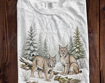 May include: A white t-shirt featuring a graphic illustration of two lynx in a snowy forest setting with evergreen trees. The shirt has a crew neckline and a Comfort Colors brand label printed on the inside of the collar area.