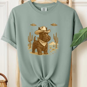 May include: A sage green t-shirt featuring a cartoon illustration of a black dog wearing a cowboy hat and bandana, set against a desert landscape with cacti and UFOs. The shirt has the words "Comfort Colors" printed on the upper back.
