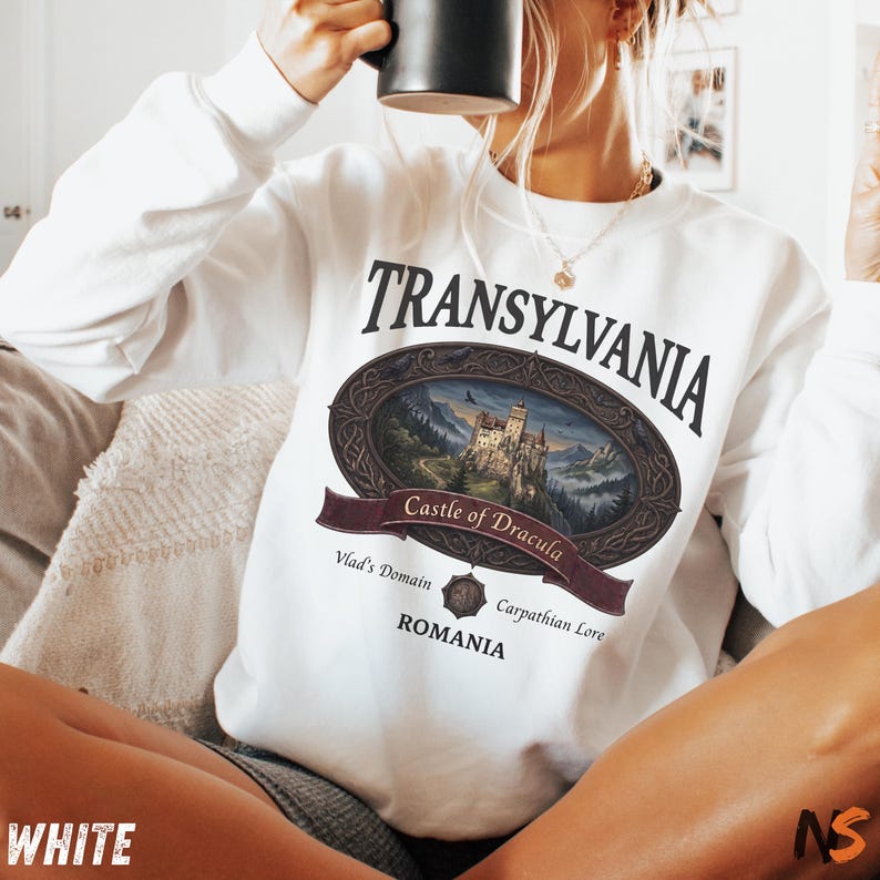Puede incluir: Una sudadera blanca de manga larga con un estampado gr&aacute;fico de un castillo dentro de un marco ovalado. El texto dice Transylvania, Castle of Dracula, Vlad's Domain, Carpathian Lore y Romania. El dise&ntilde;o incluye una banda oscura y un borde adornado.