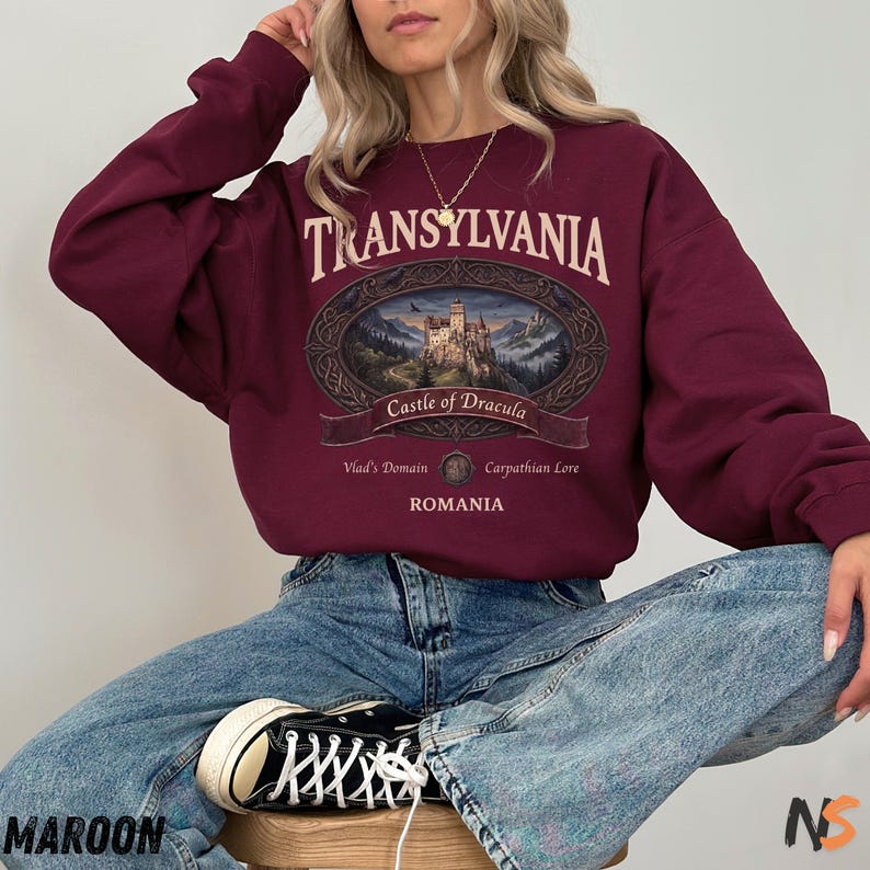 Puede incluir: Una sudadera de manga larga color granate con un estampado gr&aacute;fico que incluye el texto TRANSYLVANIA, Castle of Dracula, Vlad's Domain, Carpathian Lore y ROMANIA. La ilustraci&oacute;n central muestra un castillo en un paisaje monta&ntilde;oso.