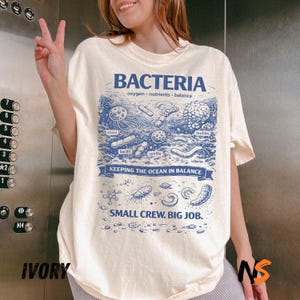 May include: An ivory t-shirt featuring a blue graphic print of various bacteria with labels including cocci, biofilm, bacilli, spirilla, and vibrio. The text reads BACTERIA, oxygen, nutrients, balance, KEEPING THE OCEAN IN BALANCE, and SMALL CREW. BIG JOB.