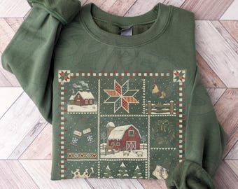 May include: An olive green crewneck sweatshirt featuring a central graphic panel with a patchwork design. The print displays a red barn, a rooster, a star, winter mittens, a wreath, and snowy landscapes in shades of red, white, and brown.