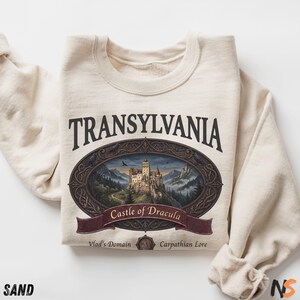 Puede incluir: Una sudadera de cuello redondo color arena con un estampado gr&aacute;fico que incluye el texto TRANSYLVANIA, Castle of Dracula, Vlad's Domain y Carpathian Lore. El dise&ntilde;o presenta un marco ovalado que rodea un castillo.