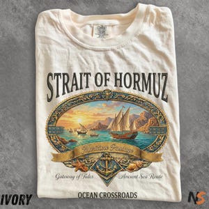 May include: Ivory-colored t-shirt featuring a detailed graphic of the Strait of Hormuz. The design includes sailing ships, a sunset scene, and text that reads "Maritime Passage" and "Ocean Crossroads." The shirt also has the text "Comfort Colors."