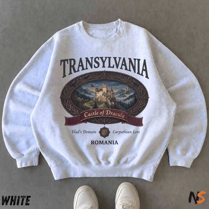 Puede incluir: Una sudadera de cuello redondo gris claro con un estampado gr&aacute;fico con el texto TRANSYLVANIA, Castle of Dracula, Vlad's Domain, Carpathian Lore y ROMANIA. La imagen central muestra un castillo en la cima de una monta&ntilde;a en un marco ovalado.