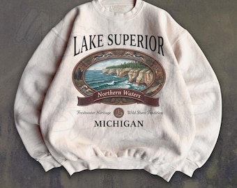 May include: A cream-colored crewneck sweatshirt featuring a graphic of Lake Superior with the text "LAKE SUPERIOR," "Northern Waters," "Freshwater Heritage," "Wild Shore Tradition," and "MICHIGAN" printed in black and brown tones.