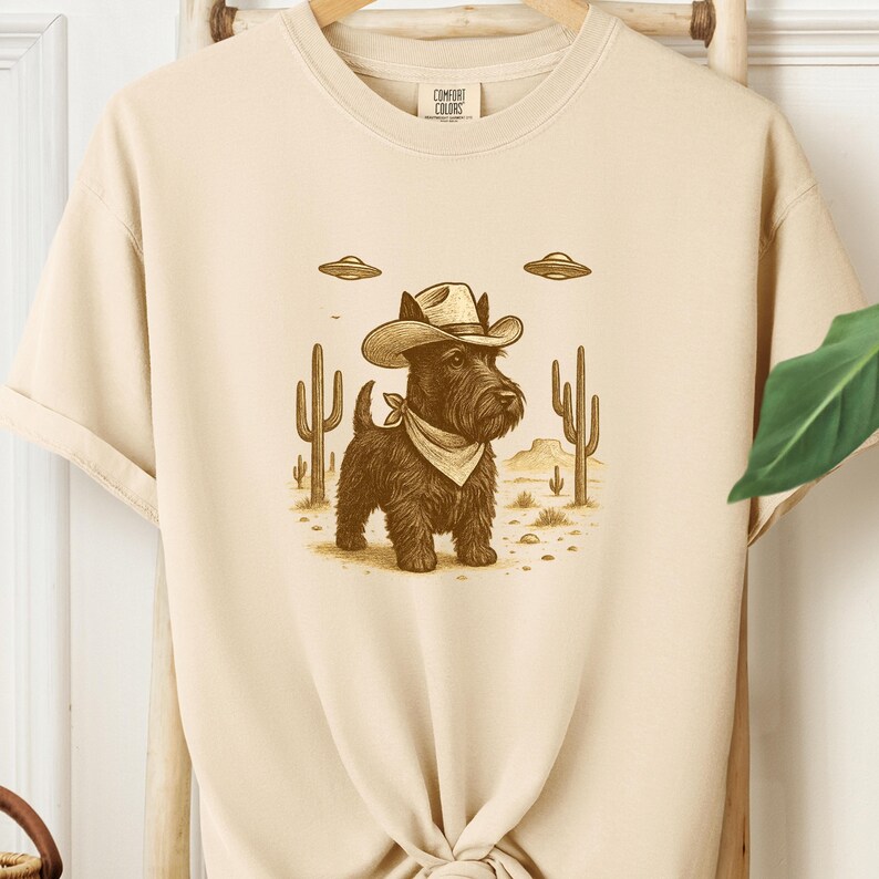 May include: A beige Comfort Colors t-shirt featuring a cartoon illustration of a black dog wearing a cowboy hat and bandana, with cacti and UFOs in the background. The shirt has a crew neck and short sleeves.
