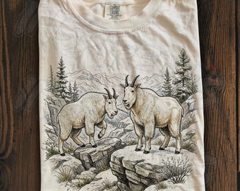 May include: An ivory-colored t-shirt featuring a detailed graphic illustration of two mountain goats standing on rocky cliffs. The scene includes pine trees and mountain peaks in the background. The shirt has a crew neckline and a Comfort Colors label.