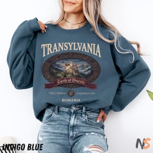 Puede incluir: Una sudadera de cuello redondo color azul &iacute;ndigo con un estampado gr&aacute;fico que incluye el texto TRANSYLVANIA, Castle of Dracula, Vlad's Domain, Carpathian Lore y ROMANIA. El dise&ntilde;o presenta un marco ovalado que rodea una ilustraci&oacute;n detallada de un castillo.
